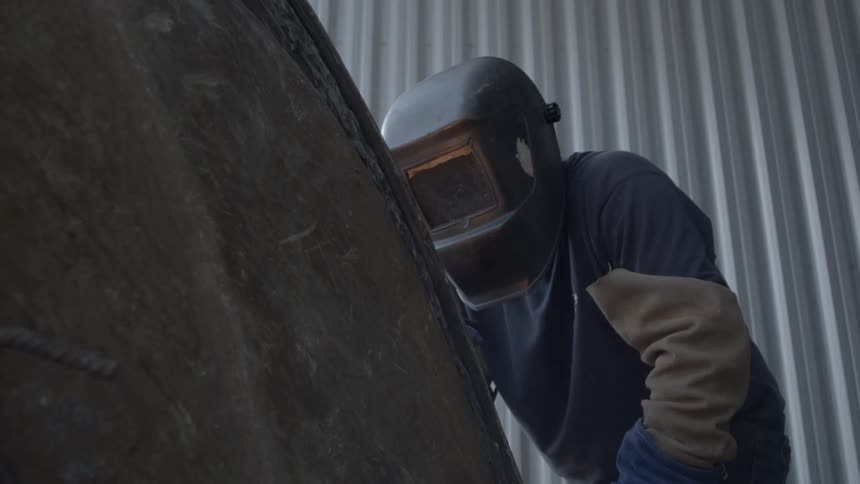 A welder in a protective mask works on a large piece of metal with sparks flying in slow motion