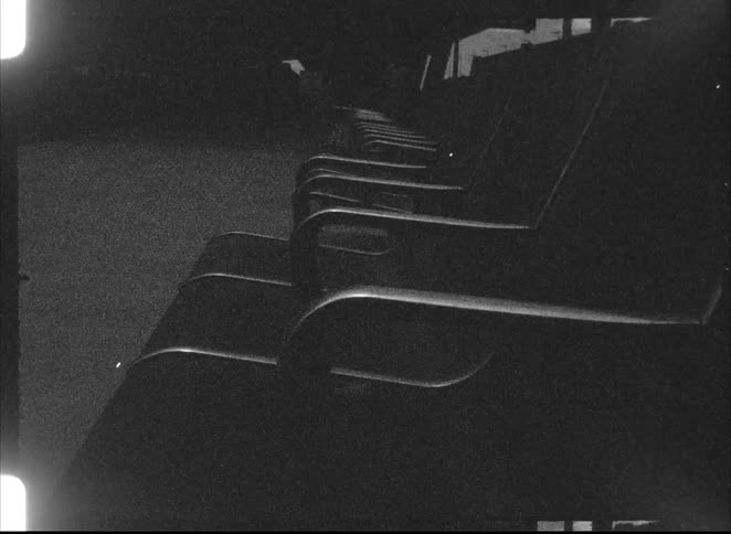 A grainy, black and white, low-angle shot captures a row of empty seats in a dark, atmospheric theater