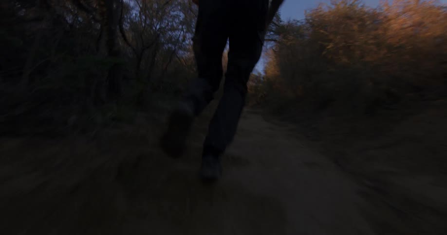 A low-angle shot follows a person's feet as they run along a dark trail through dense brush at twilight