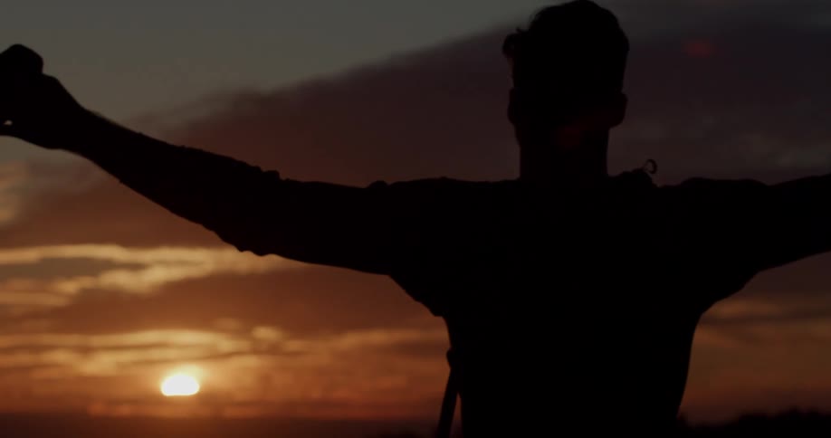 A silhouette of a person raises their arms wide against a beautiful sunset sky
