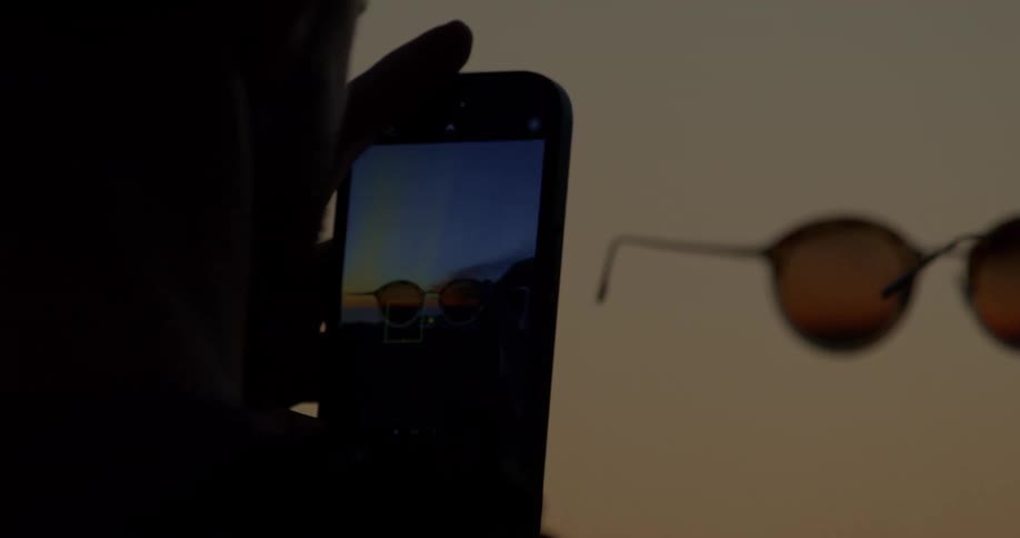 Silhouette of a person taking a creative photo of a beautiful sunset reflected in sunglasses using a smartphone
