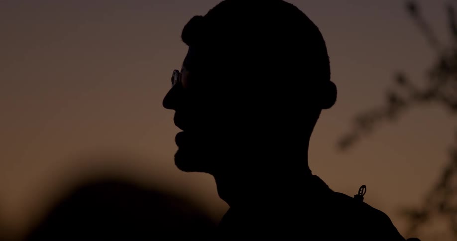 A close-up silhouette of a man with glasses and a beanie looking contemplatively at the serene dusk sky