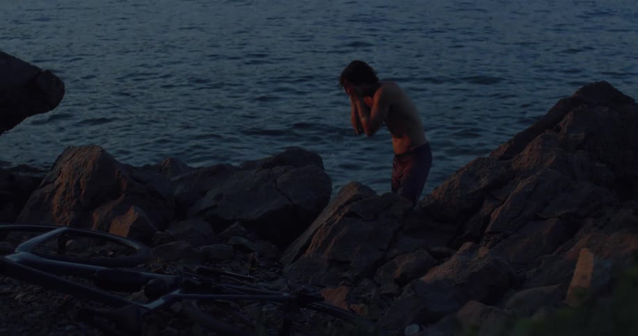 A young man splashes water on his face to cool down on a rocky seashore at dusk in slow motion