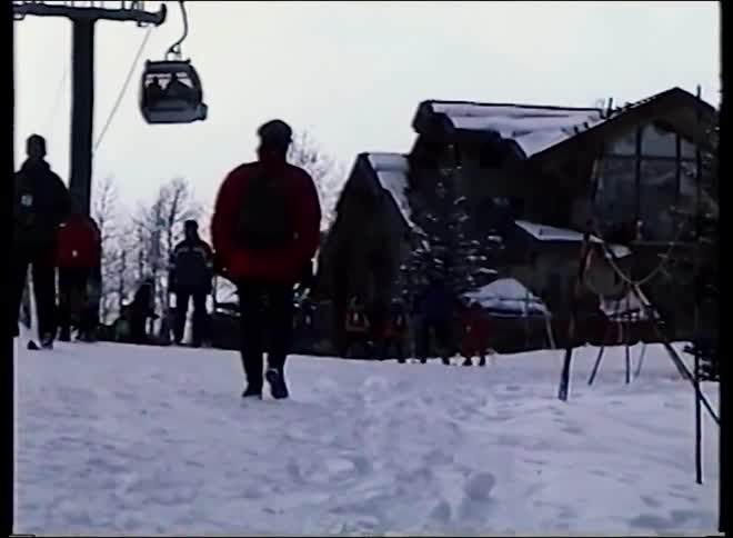 Vintage footage shows people at a ski resort with gondolas passing overhead as a person in a red jacket walks up the snowy hill