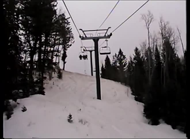 A vintage POV shot from a chairlift moving up a snowy mountain slope on an overcast day