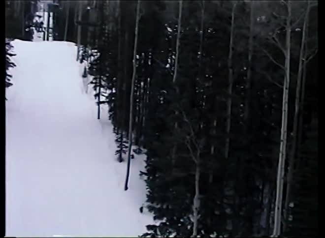 POV from a moving ski lift passing over a large wooden chalet nestled in a snowy, wooded mountain landscape