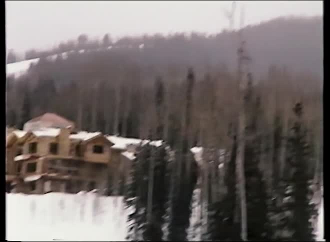 A vintage-style shot shows people riding a ski lift up a snowy mountain past a large lodge in the woods