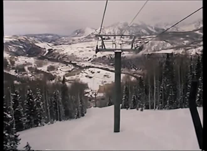 A first-person view from a chairlift traveling down a snowy mountain slope with a vast winter landscape in the background