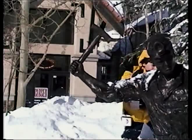A little boy in a yellow winter jacket playfully throws snow at and poses with a bronze miner statue in a snowy village