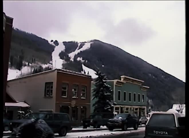 Vintage footage shows a snowy street in a classic ski resort town, with a large mountain visible in the background