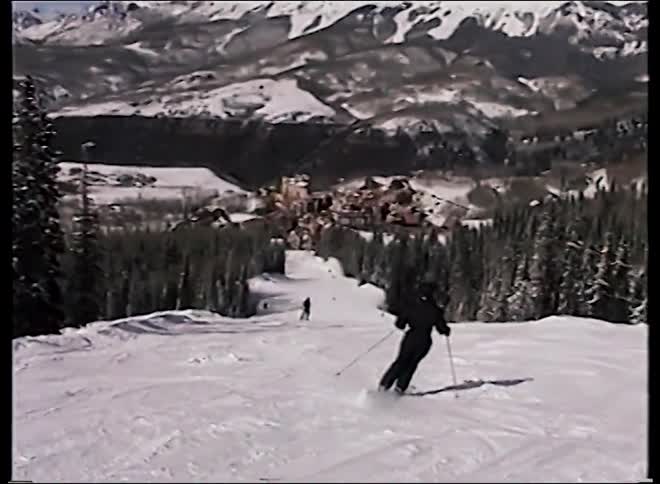 Vintage home video footage follows a skier down a sunny slope toward a picturesque mountain village and ski resort