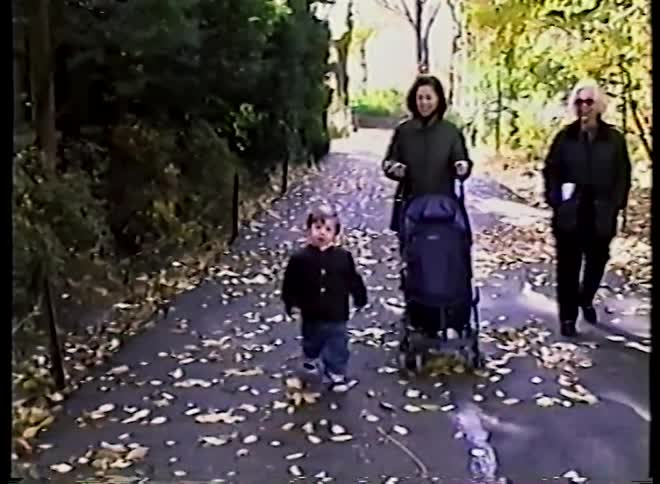 Slow motion vintage footage shows a happy toddler walking down a leaf-strewn path with his mother and grandmother on a sunny autumn day