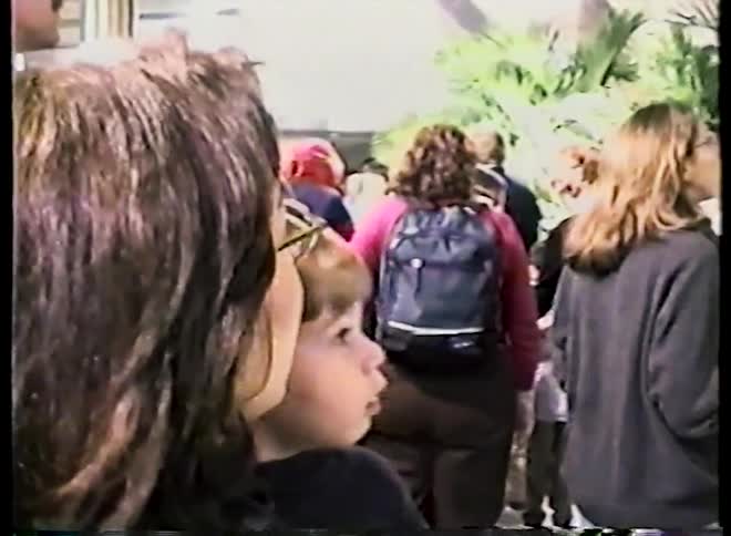 Vintage home video captures a mother smiling while holding her curious son in a crowded public space