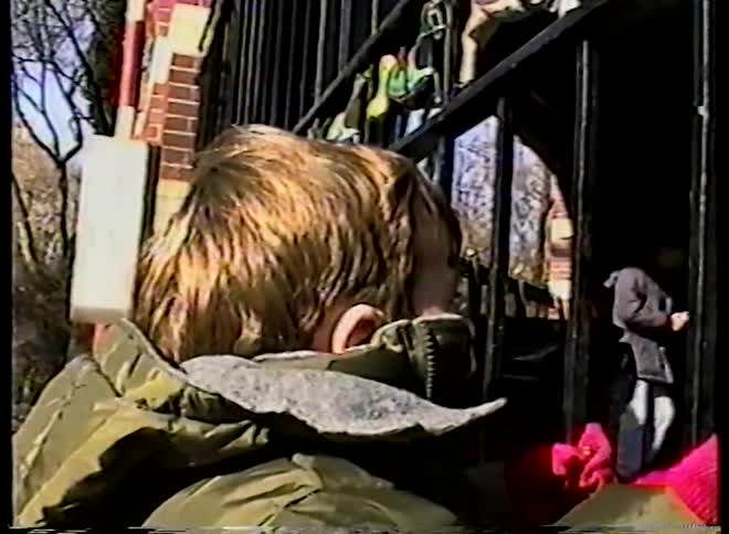 Vintage VHS footage captures a small child in a winter coat watching a carousel from behind a fence
