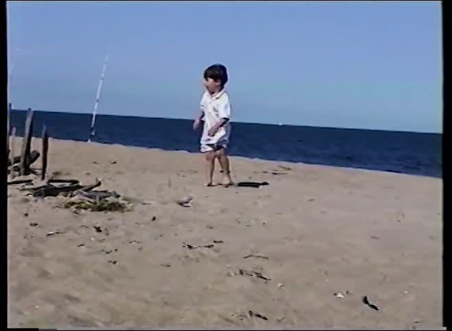 Vintage home video footage shows a young boy playfully dancing on a sunny beach by the ocean