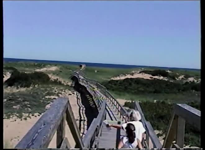 Vintage home video shows a family walking along a wooden boardwalk over sand dunes towards the ocean on a sunny day