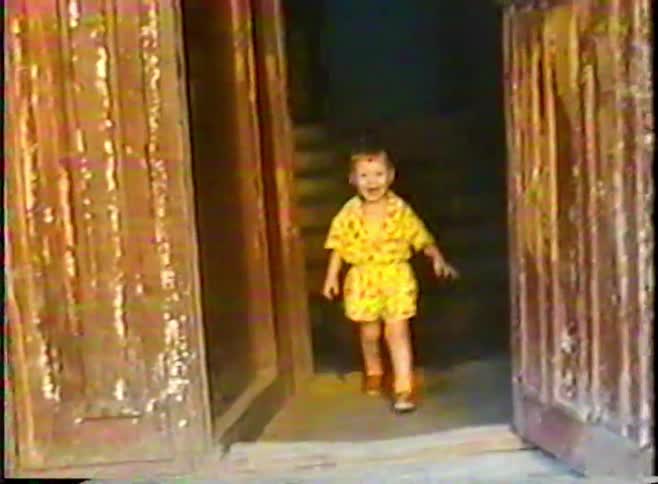 Vintage, distorted VHS footage shows a happy toddler in a yellow outfit walking out of a dark doorway