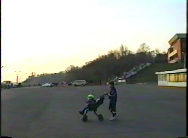 Vintage footage captures a boy on rollerblades carefully pushing his younger sibling in a three-wheeled stroller across a parking lot at sunset