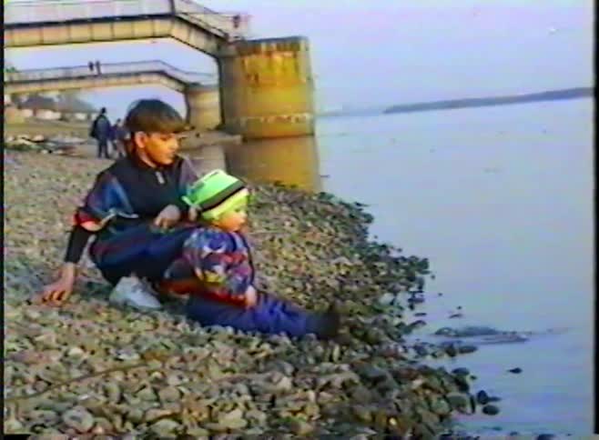 Vintage 90s footage of two young brothers sitting together on a rocky riverbank next to a bridge