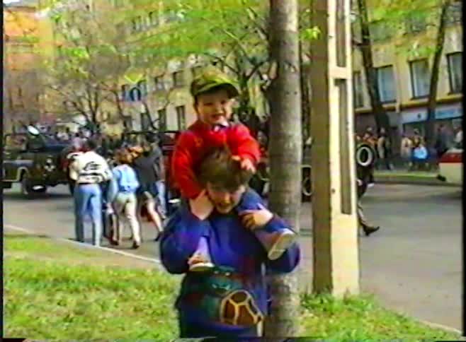 A vintage home video shows a young boy giving his sibling a shoulder ride to get a better view of a passing military parade on a city street