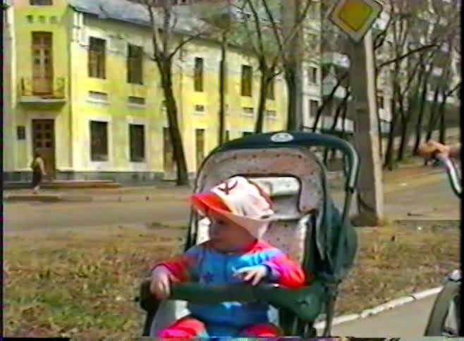 A young boy with his bicycle stands by a baby in a stroller wearing a hat with a Soviet hammer and sickle symbol on a city sidewalk