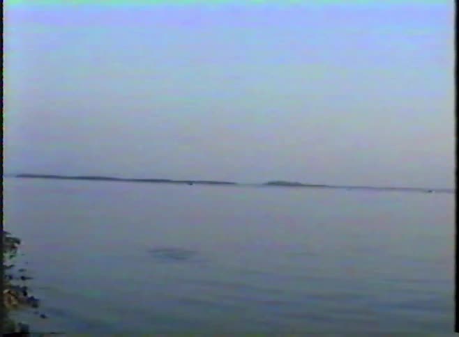 Archival footage captures two children launching a toy sailboat from a rocky shore onto a vast, calm lake