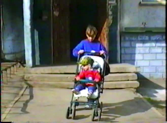 An older brother pushes his toddler sibling in a stroller away from their apartment building entrance in this retro home video