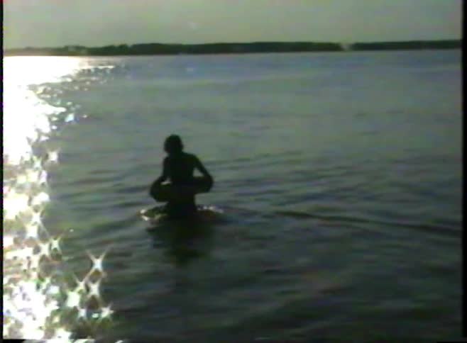A silhouetted boy with an inner tube wades out into a sparkling lake in this vintage footage