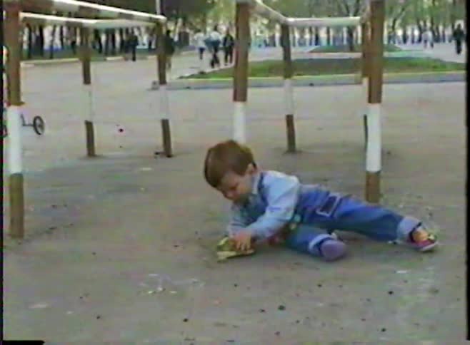 A vintage home video shows a little boy in overalls hanging from a bar and then playing with his toy car in the dirt at a park