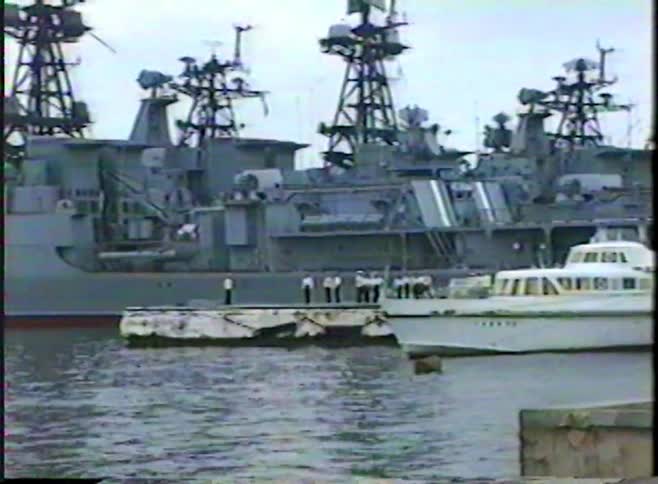 Soviet-era sailors stand on a pier beside a large naval warship docked in a harbor
