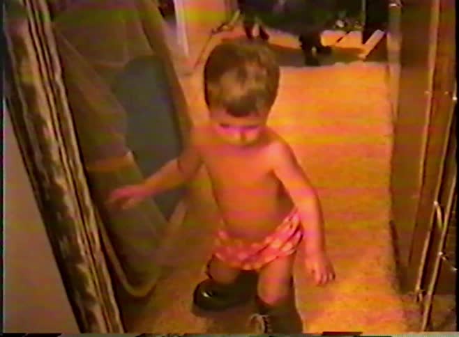 Vintage home video shows a small child in just a diaper trying to walk in a pair of oversized boots