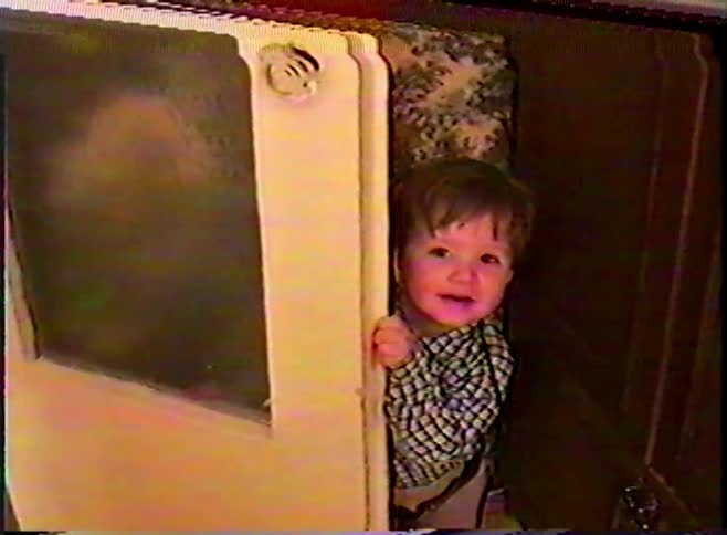 A cute little boy plays peek-a-boo from behind a door and smiles in a retro home movie