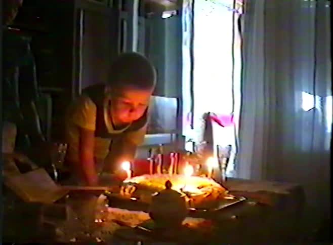 A young boy struggles to blow out the candles on his birthday cake in a nostalgic vintage home movie