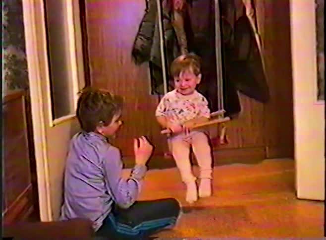 A heartwarming vintage VHS home video of a boy sitting on the floor watching his happy toddler sibling on an indoor swing