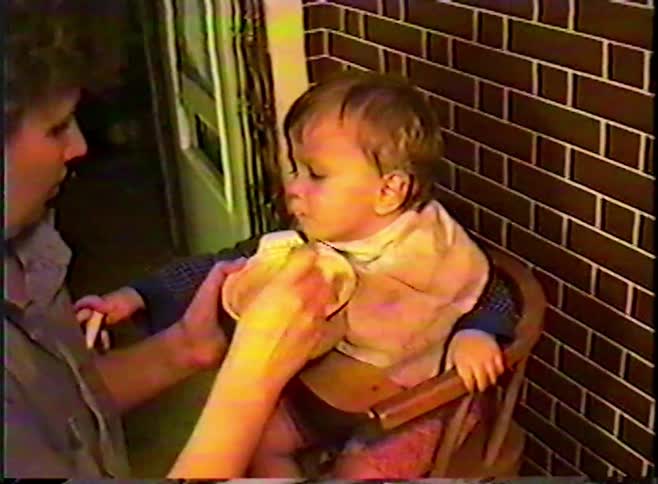 A nostalgic VHS home movie from the 1980s shows a mother spoon-feeding her baby in a wooden high chair
