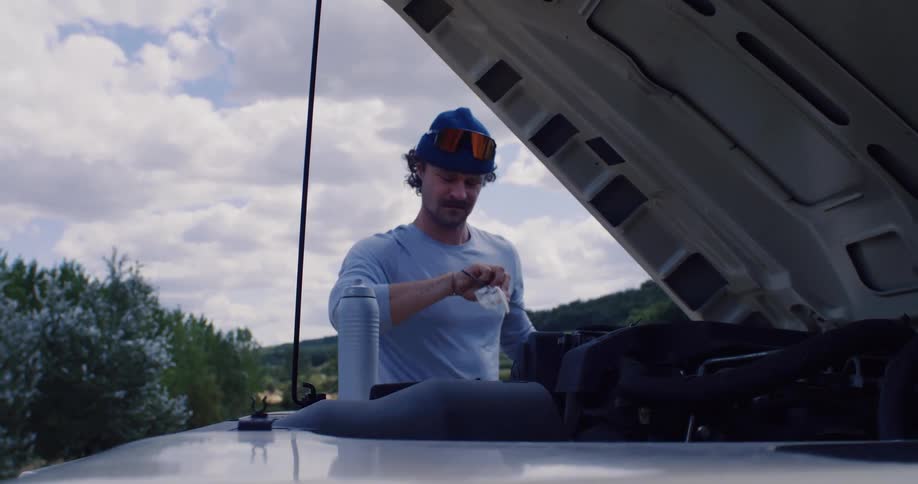 A man wearing a blue beanie and sunglasses checks the oil level of his truck with the hood propped open on a partly cloudy day