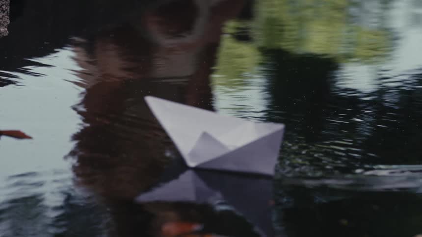 The reflection of a person in a tranquil pond launches a paper boat that gently floats away