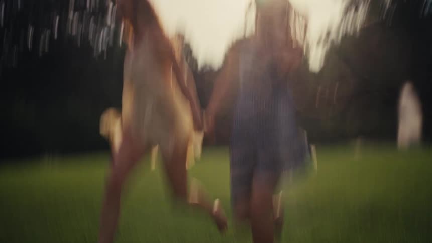 Two joyful young women in dresses hold hands and run freely and barefoot across a sun-drenched lawn in a beautiful garden at golden hour
