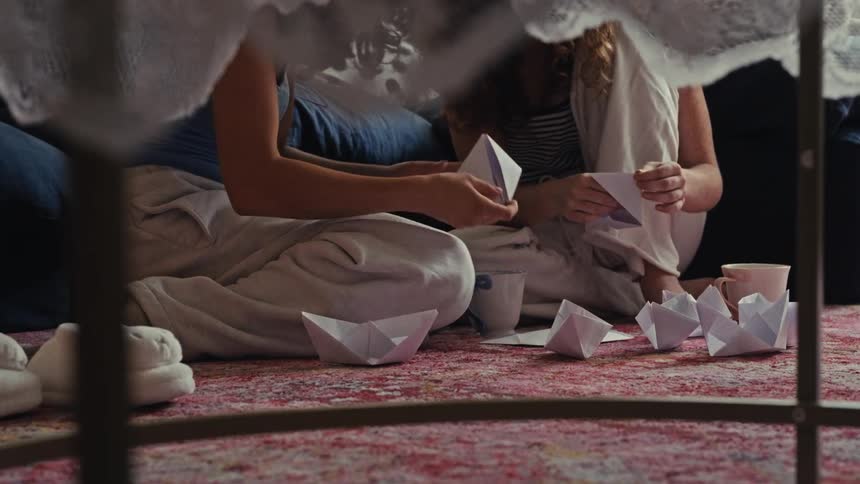 Two friends bond while making a fleet of origami paper boats inside a cozy homemade blanket fort