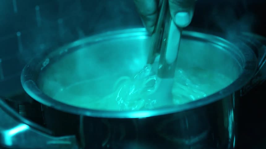 A chef stirs spaghetti in a pot of boiling water under moody cyan lighting in a dark kitchen