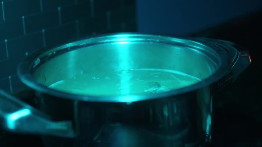 A chef uses tongs to stir something in a boiling pot on a stove under a mysterious cyan light