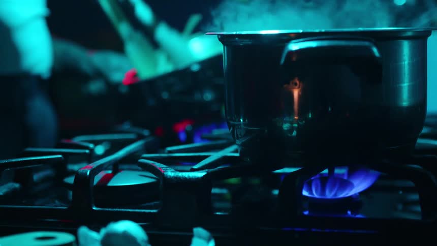 Close up on a steaming pot on a gas burner as a chef prepares a meal in a dark, atmospheric kitchen