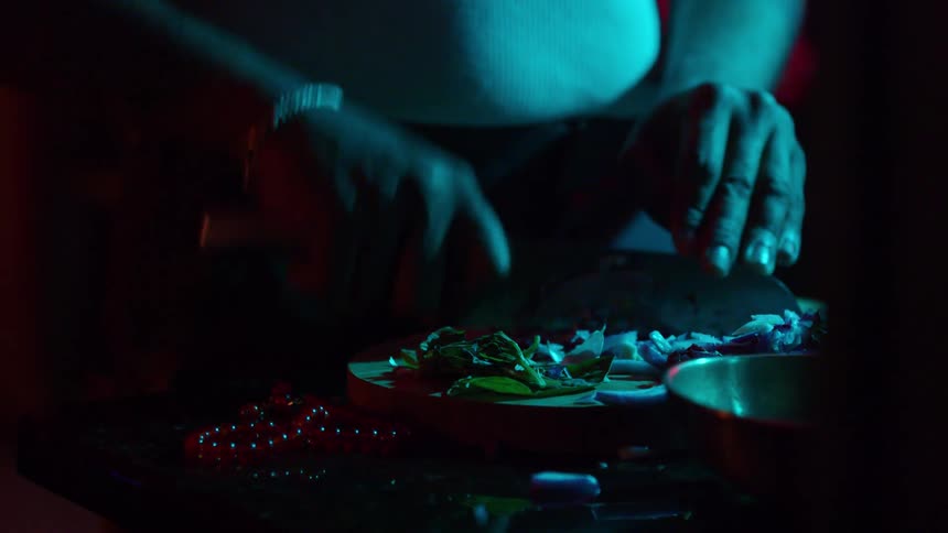 Under atmospheric blue and red light, a person carefully chops vegetables on a wooden cutting board