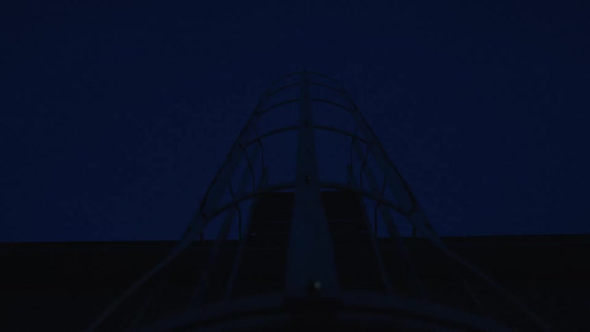 A low angle, stationary shot of a silhouetted industrial metal ladder with a safety cage against the deep blue night sky