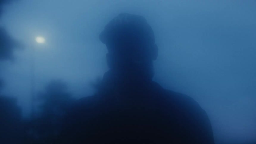 A mysterious silhouette of a person smokes a cigarette in dense blue fog under a street lamp at night