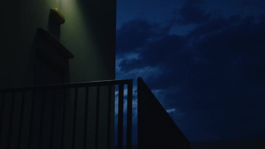 An atmospheric view from a dark, modern balcony at night shows a single light illuminating a wall as clouds move across the deep blue sky