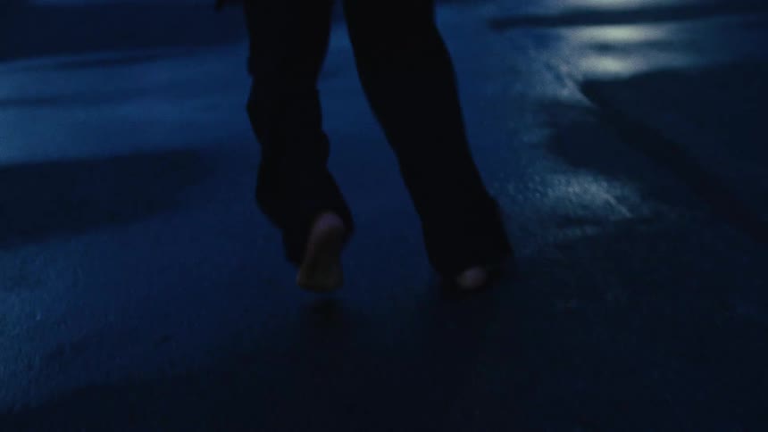 A silhouetted figure moves erratically barefoot on a wet street at dusk, falling and crawling like a creature