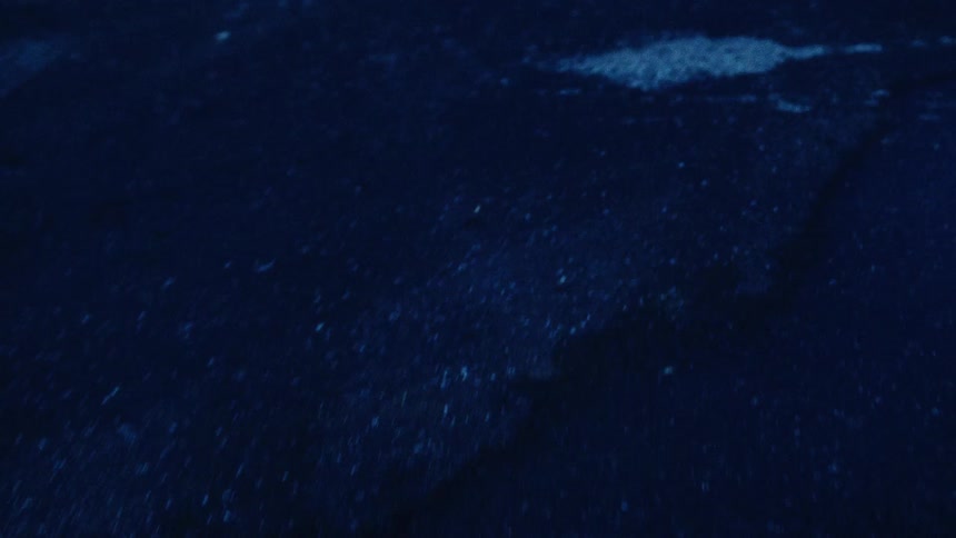 A dramatic backward tracking shot captures raindrops splashing onto wet asphalt on a dark, moody night