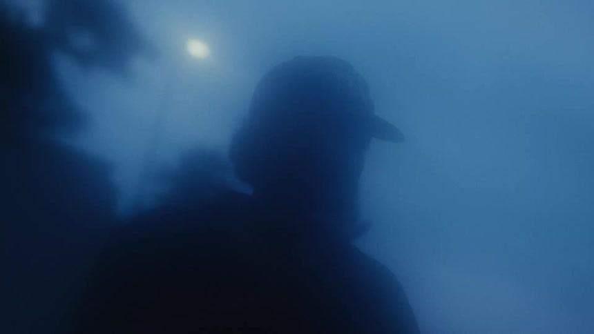 A mysterious silhouette of a person wearing a cap is obscured by thick blue fog at night