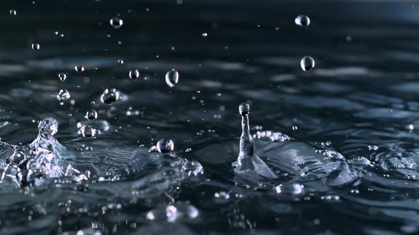 Super slow motion macro shot of water droplets falling and splashing onto a dark, reflective surface, creating beautiful ripples